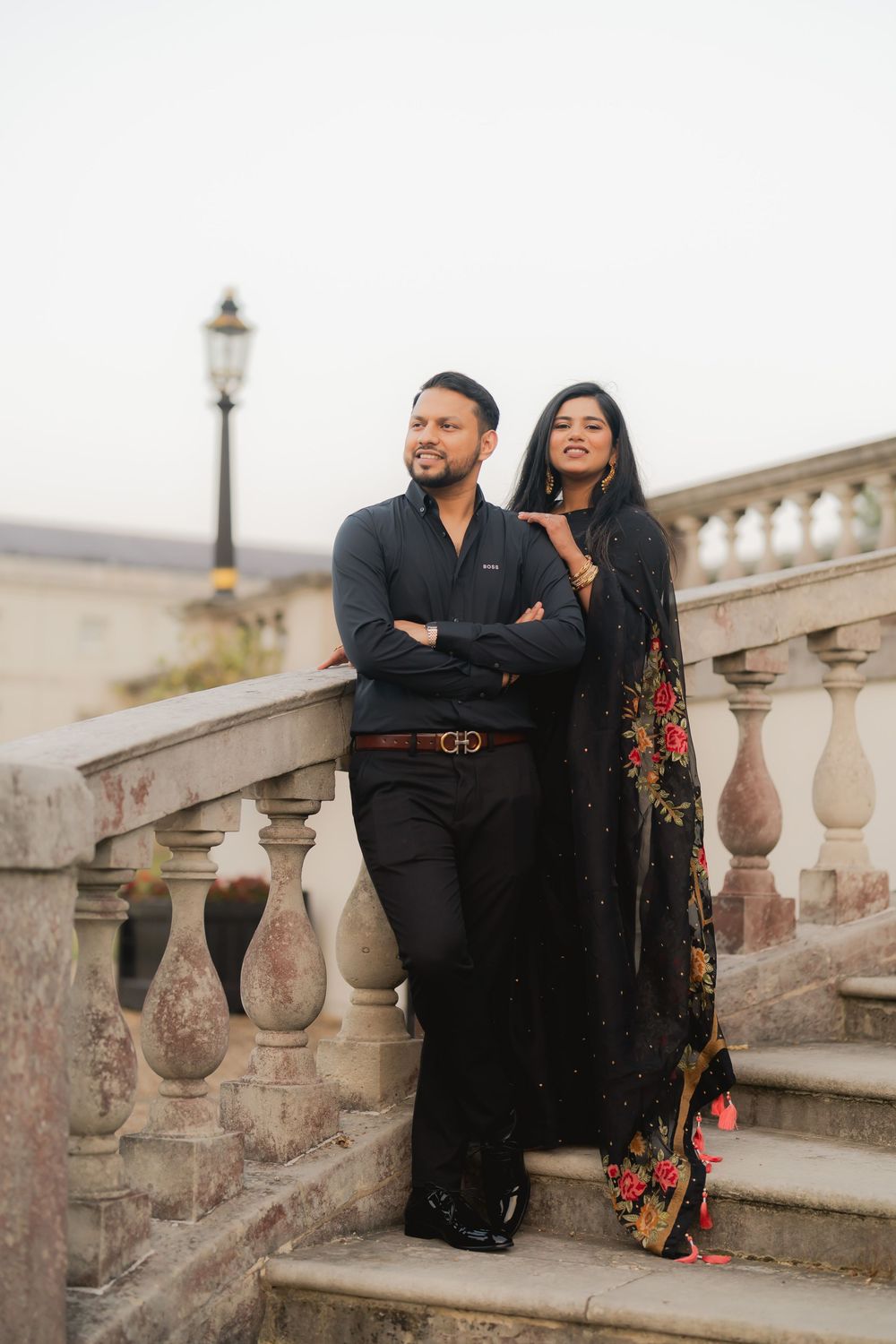 Couple in elegant black attire posing on ornate stone staircase with balustrade, woman in floral saree, man in dress shirt.