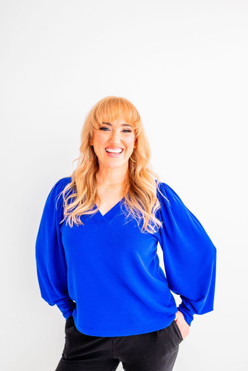 Smiling woman with long blonde hair wearing a royal blue V-neck blouse with puff sleeves, posing against a white background.