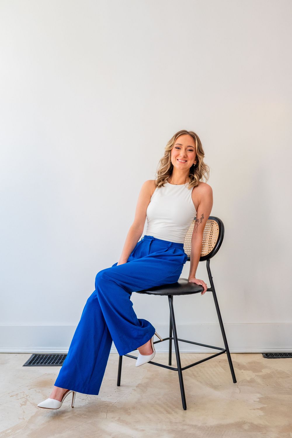 Woman in white top and royal blue wide-leg pants sitting on black bar stool against white wall.