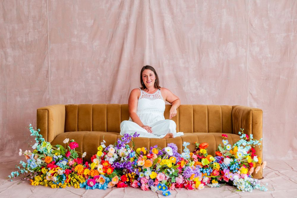 Woman in white dress sitting on gold velvet sofa surrounded by colorful flowers against pink backdrop.