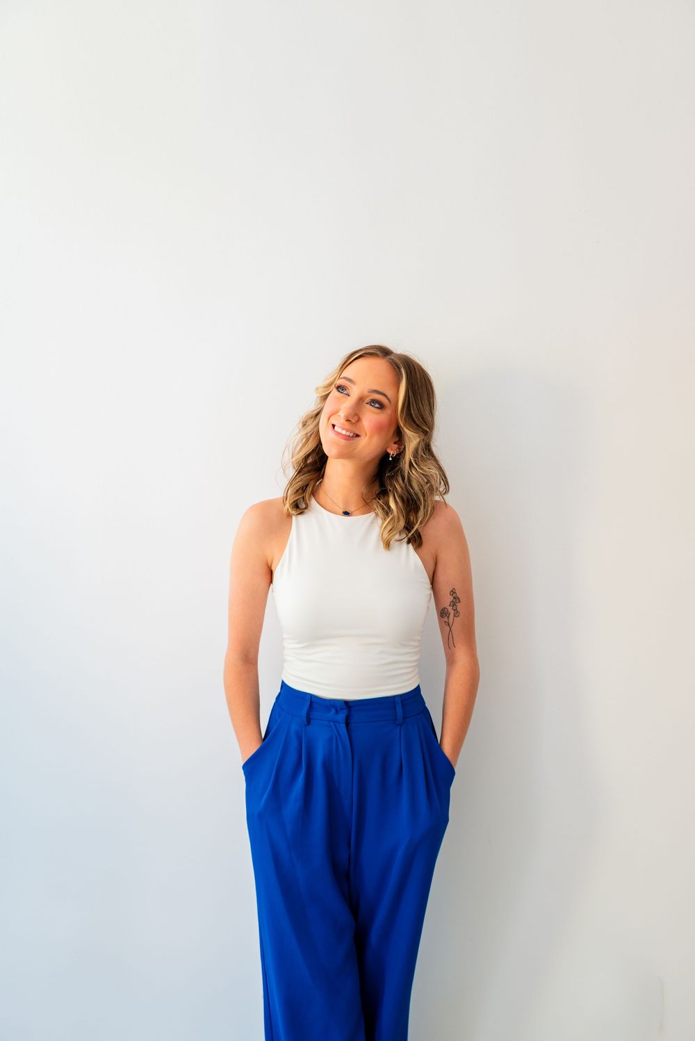 Smiling blonde woman in white tank top and royal blue wide-leg pants posing against a white wall.
