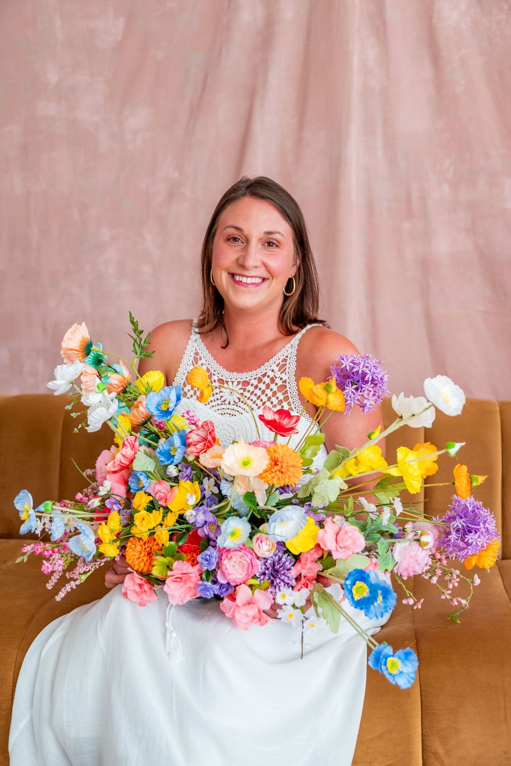 Smiling woman in white dress holding a large colorful floral bouquet while seated on a velvet sofa.
