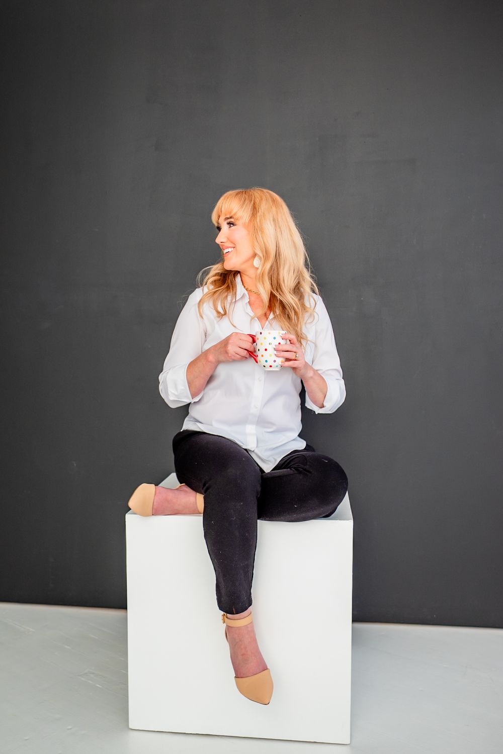 Blonde woman in white shirt and black pants sitting on white cube holding polka dot mug against gray background.