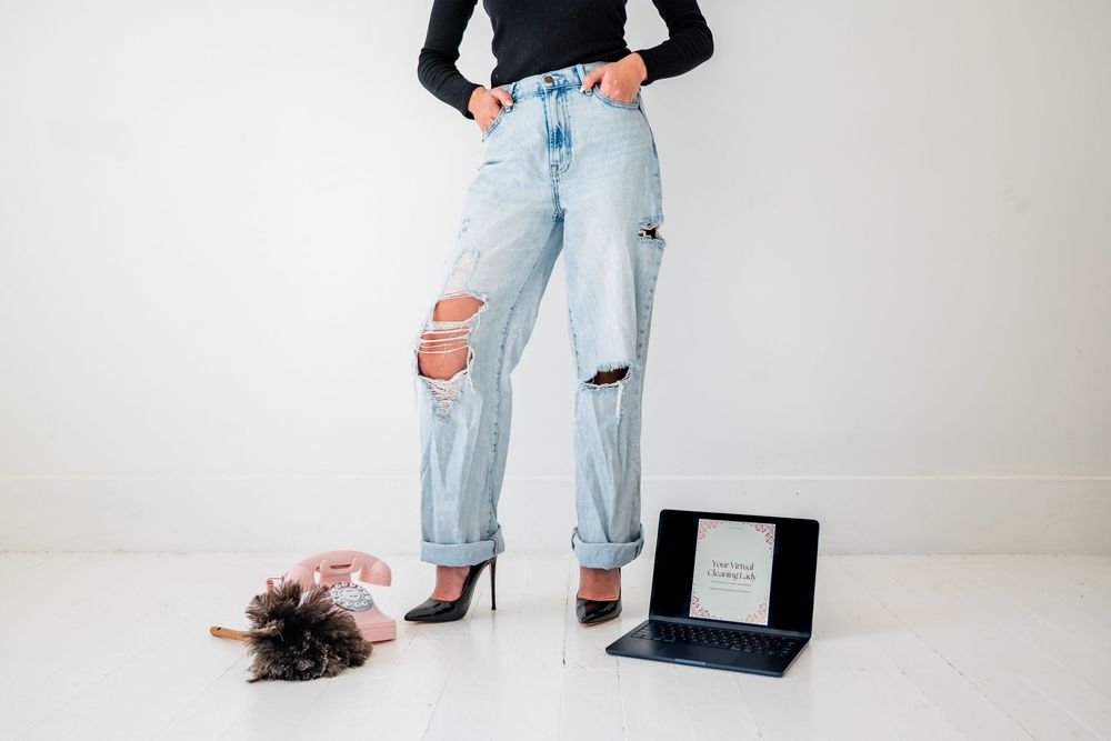 Woman in ripped jeans and black top standing next to a laptop and feather duster on white floor.