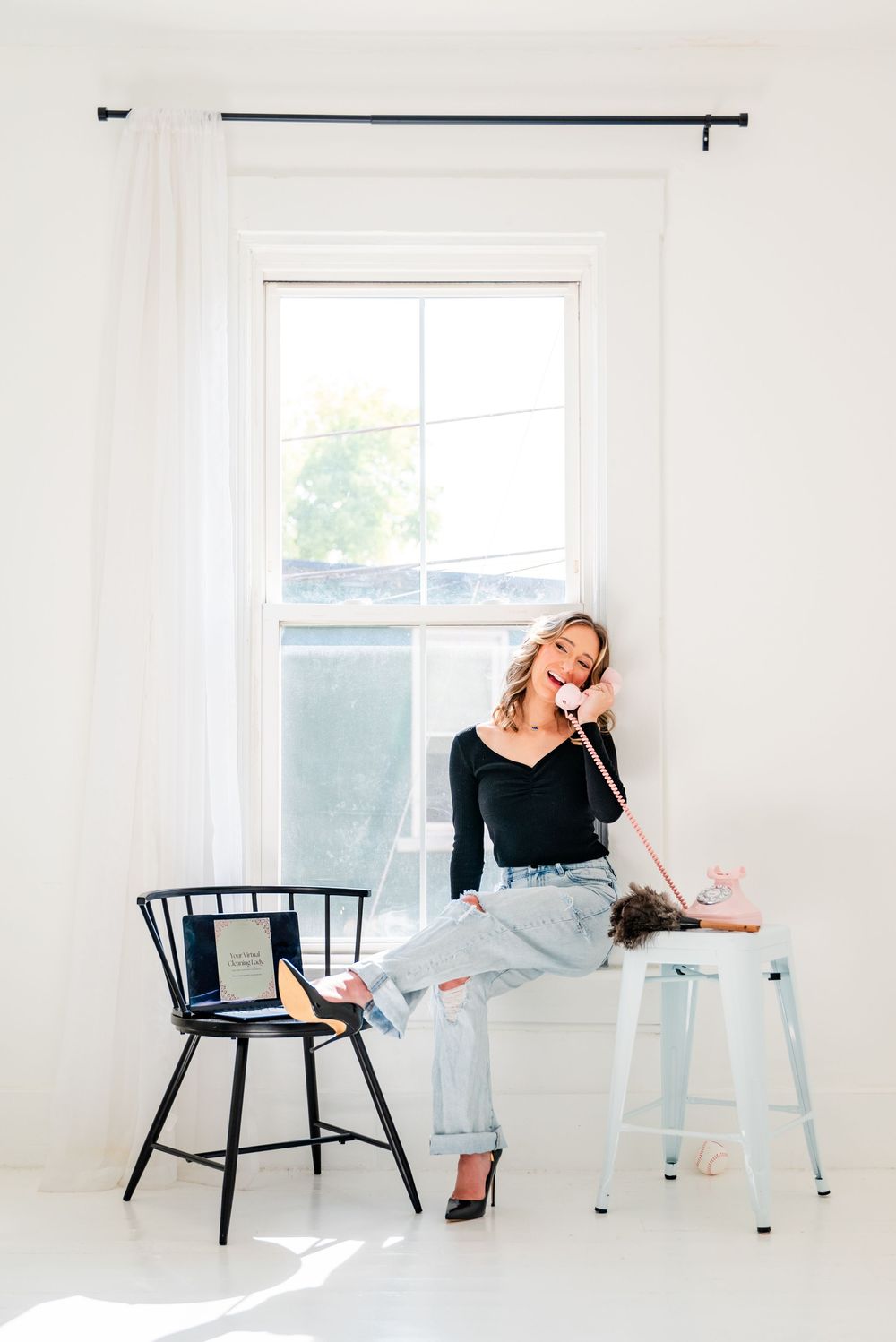 Woman in black top and jeans sitting on stool, talking on vintage pink phone in bright white room.
