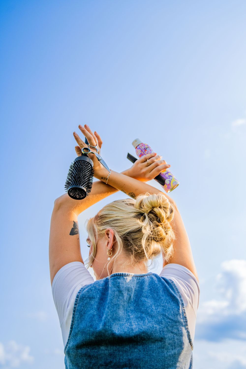 Blonde woman seen from behind holding hair brush and spray bottle up against blue sky outdoors.