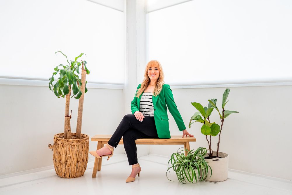 Woman in green blazer sitting on wooden bench surrounded by tropical plants in bright white room.