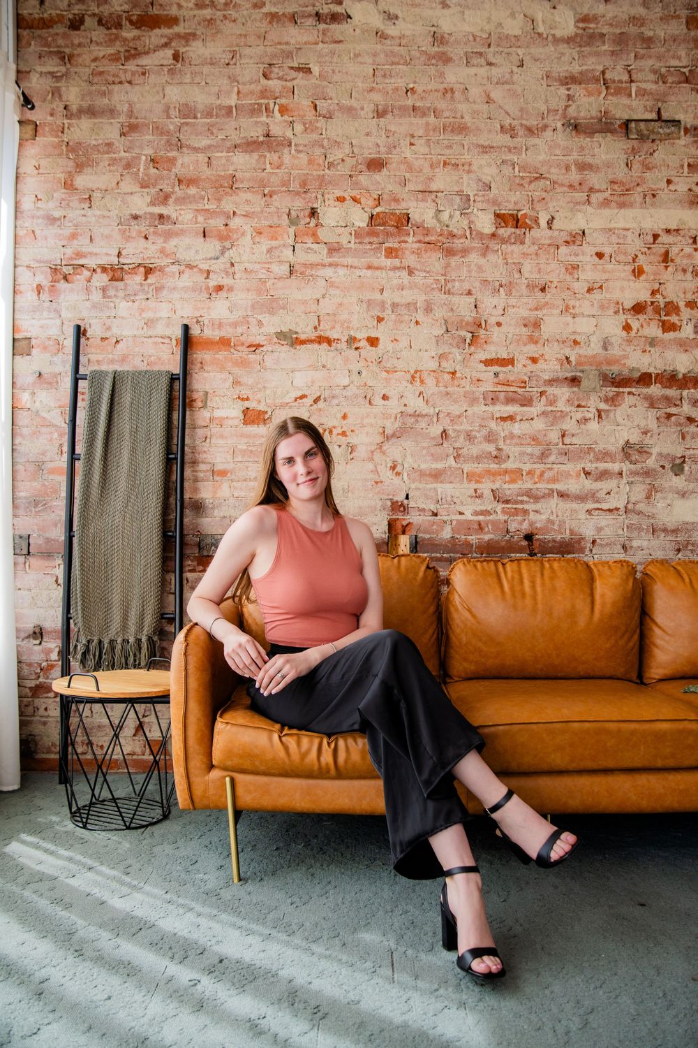 Woman in coral top and black pants sits on tan leather sofa against exposed brick wall in modern loft space.