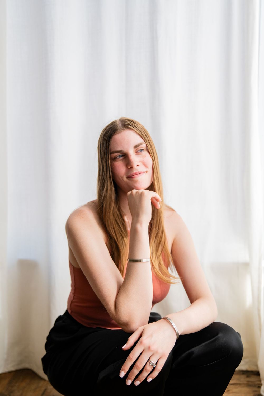 Young woman in pink top and black pants sitting cross-legged on floor, resting chin on hand, smiling softly.