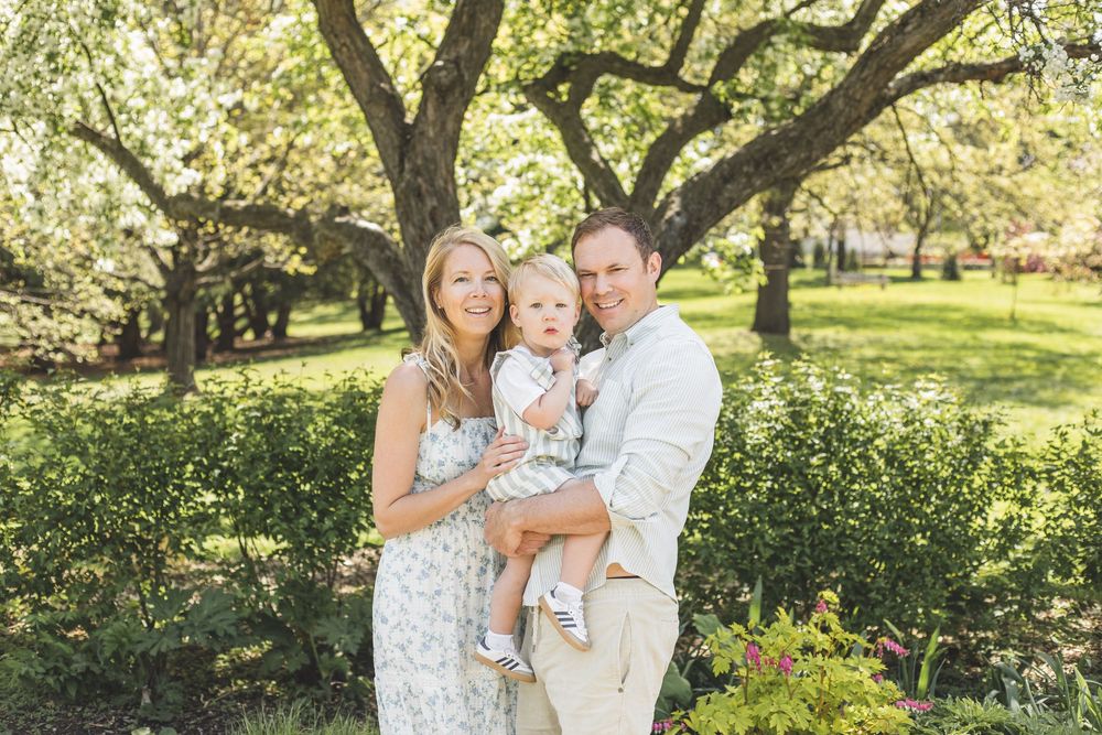 Young couple holding baby outdoors under lush green trees in bright natural light.