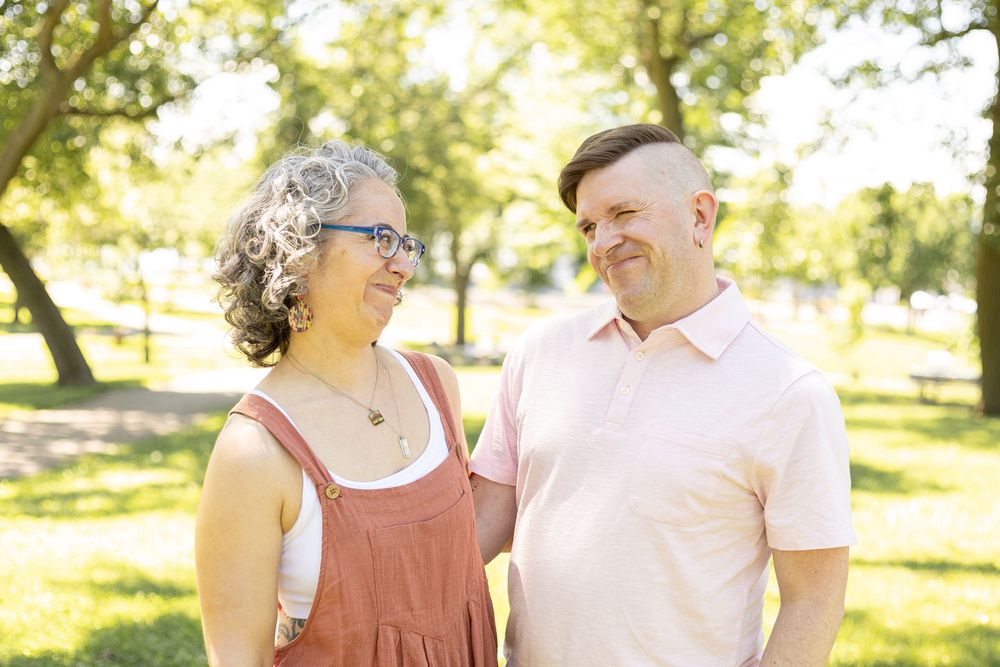Smiling couple standing together in a sunny green park, woman with curly gray hair wearing red overalls.