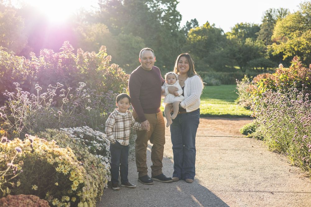 Family of four poses on a garden path surrounded by colorful autumn flowers in warm sunlight.