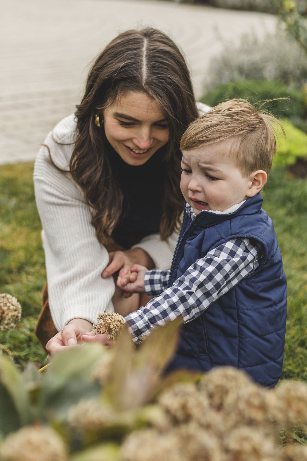 A smiling mother crouches beside her crying toddler boy in a garden, gently holding his hand outdoors.