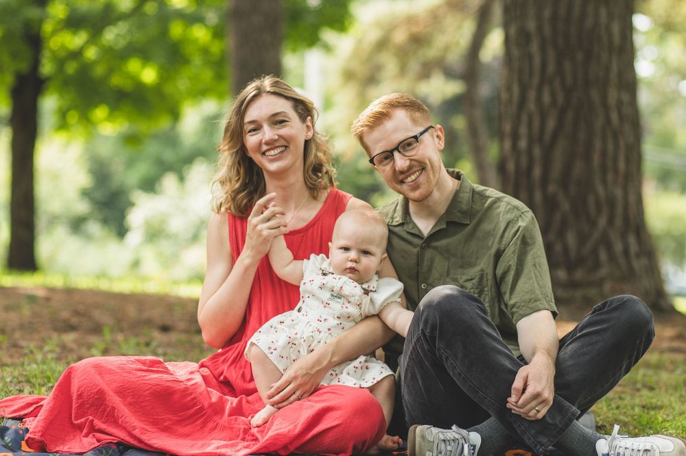 Happy young family with baby sitting on grass in a lush green park, smiling outdoors together.