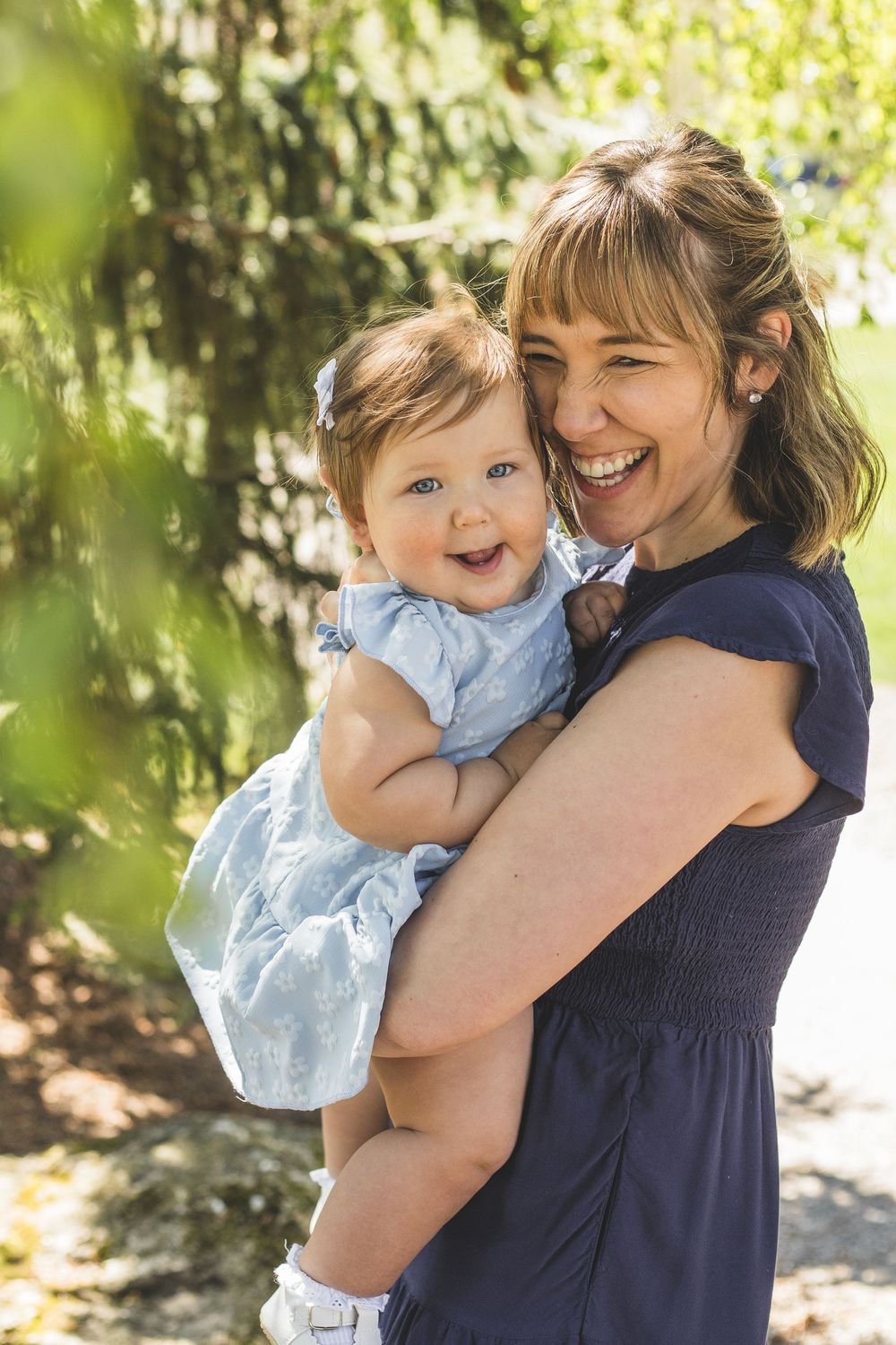 A laughing mother holds her smiling baby girl in a blue dress outdoors in a sunlit green park setting.
