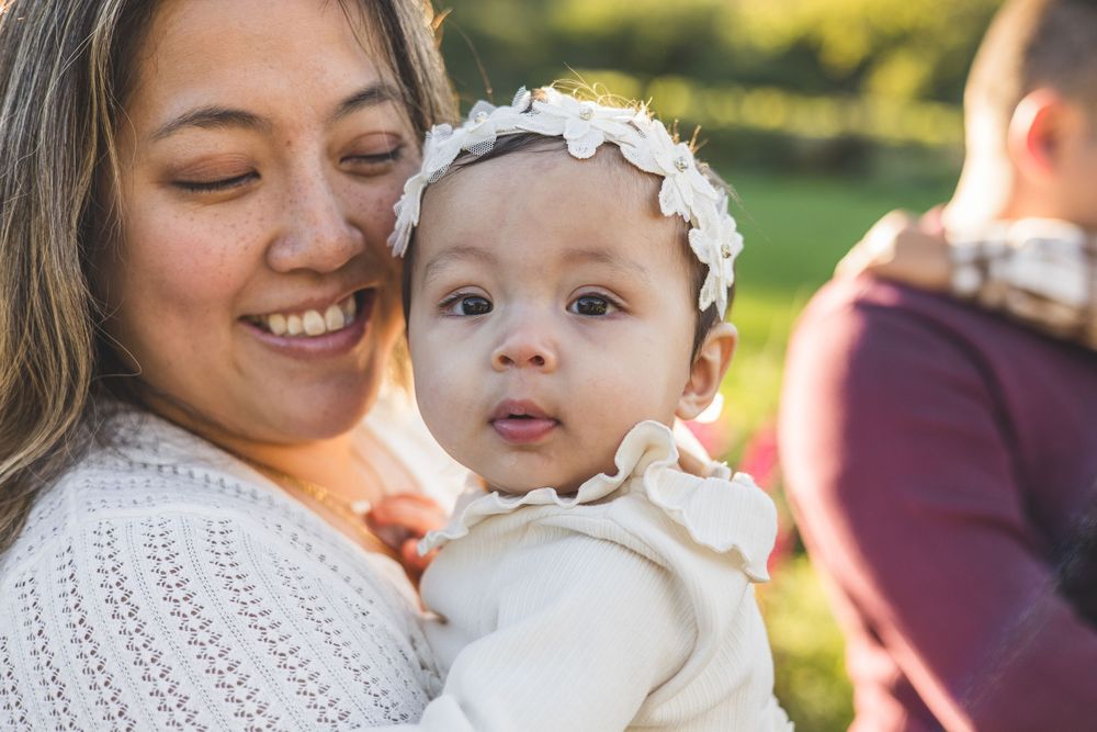 A smiling mother holds her adorable baby girl wearing a white floral headband outdoors in a sunny green park.