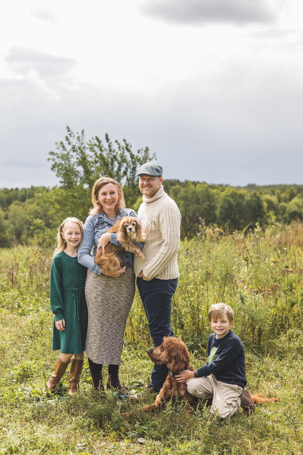 Family of four with two dogs posing in a green meadow, smiling outdoors on a cloudy day.
