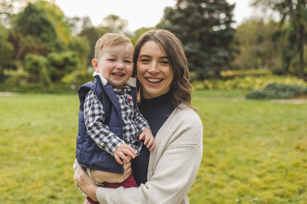Smiling mother holding happy toddler boy outdoors in lush green park setting during autumn season.