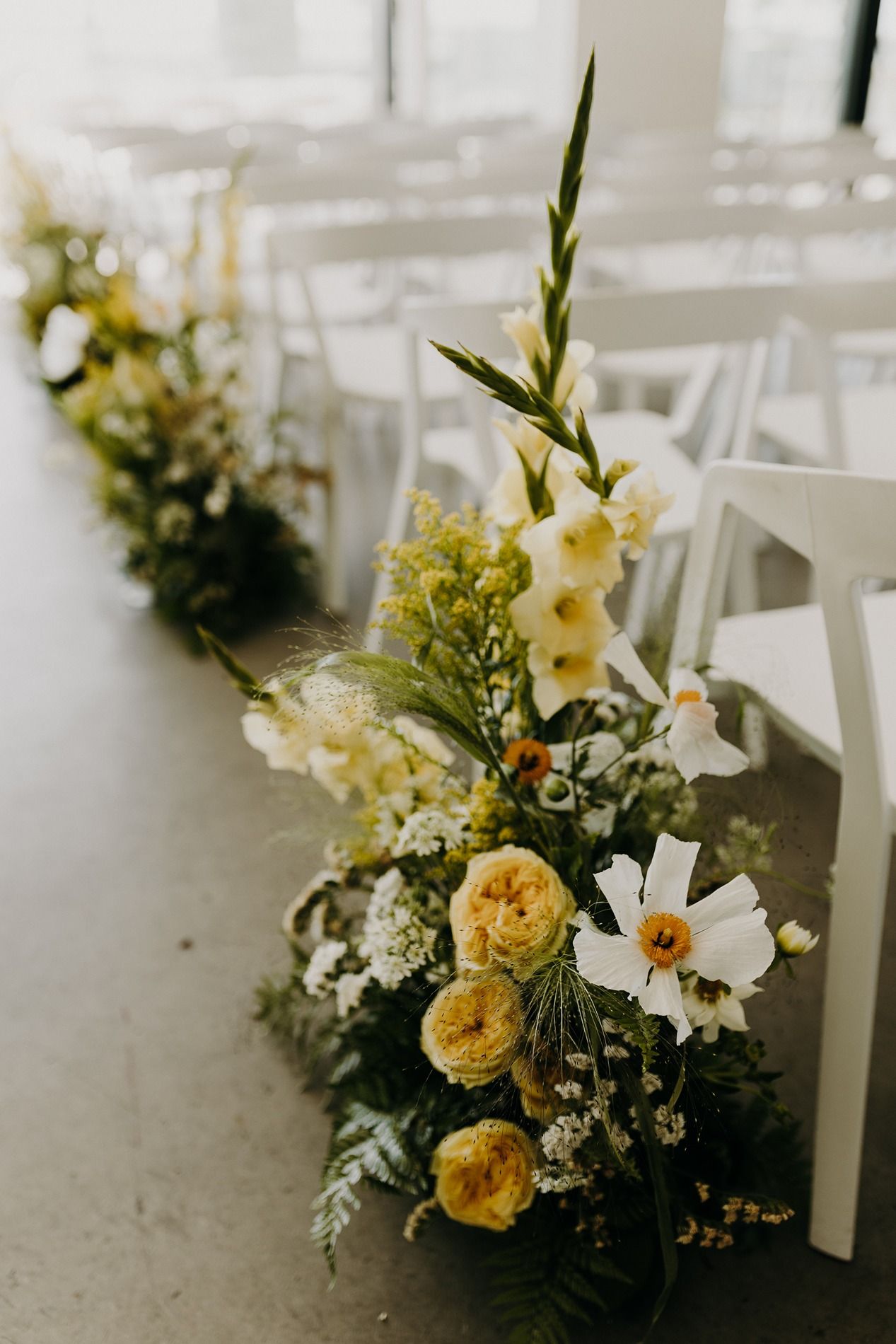 Yellow and white floral wedding aisle arrangement with roses, gladiolus, and greenery beside white chairs at The Lane.