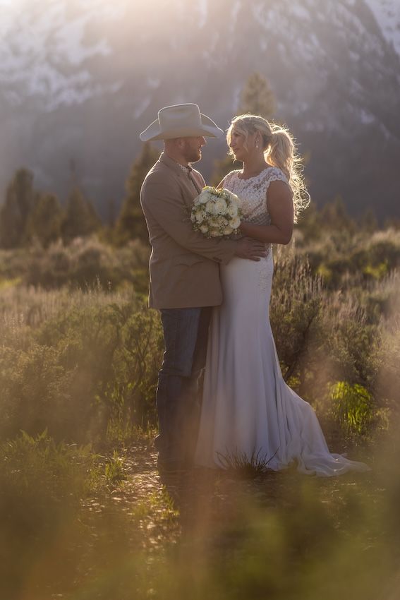 Bride and groom embrace at golden hour in a mountain meadow, she holds white bouquet, he wears cowboy hat and tan suit.