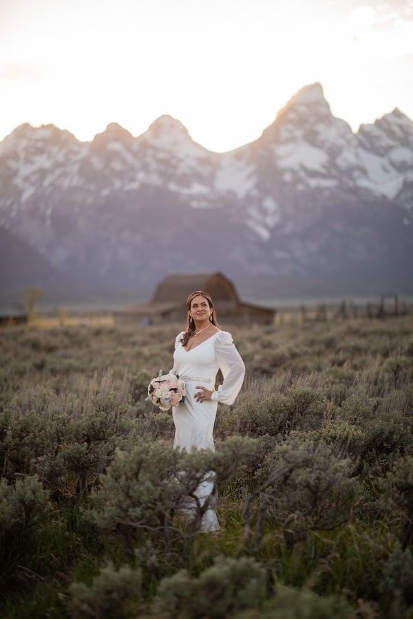 Bride in white long-sleeve gown holds bouquet in sagebrush field with Grand Teton mountains at sunset.