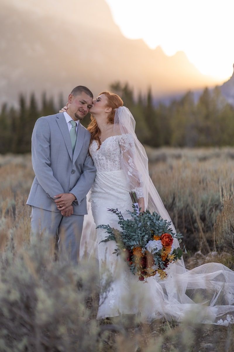 Bride and groom embrace at golden hour with mountain backdrop, bride holding colorful bouquet in white lace gown and veil.