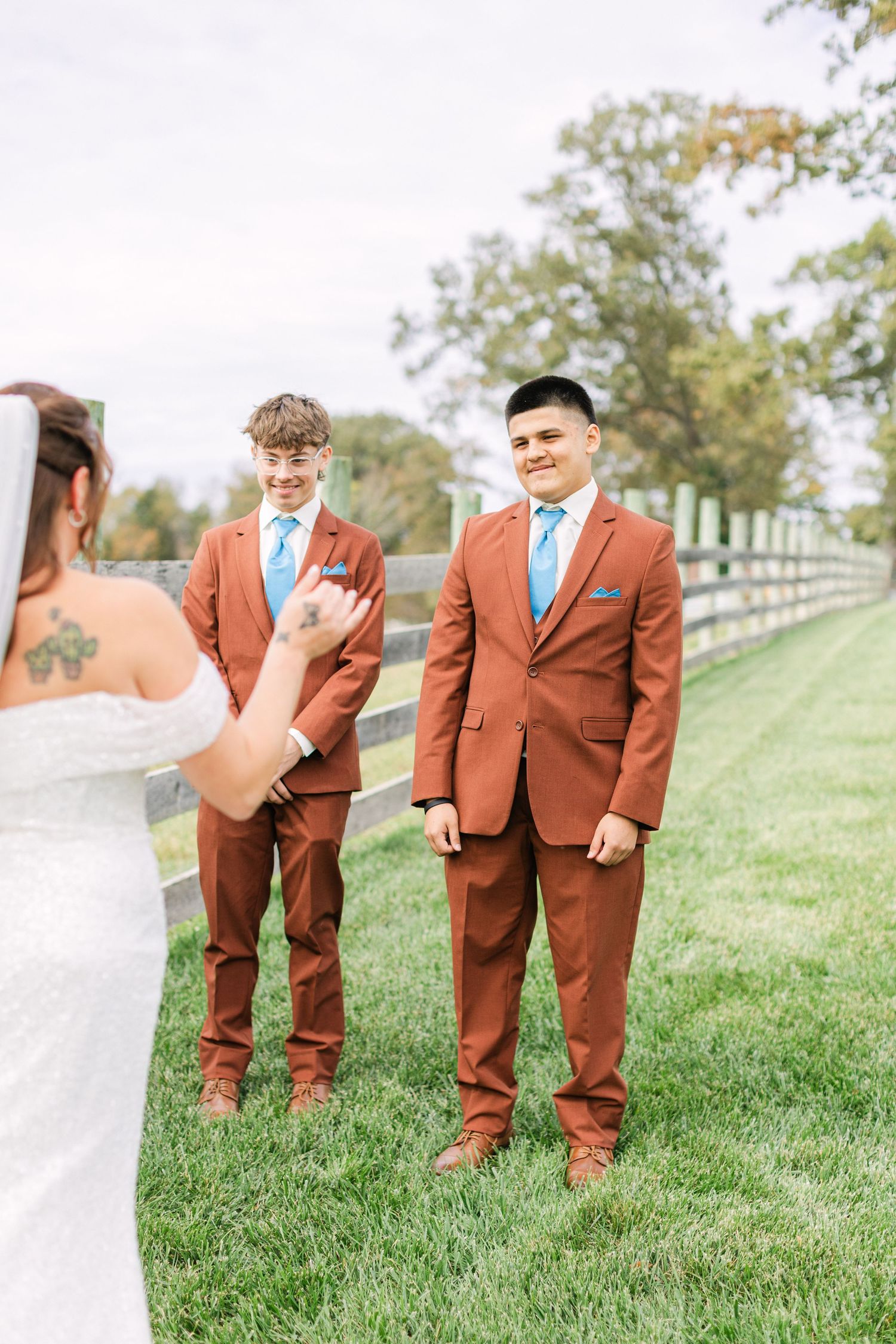 Two groomsmen in rust-colored suits with blue ties smile at a bride during an outdoor wedding ceremony.