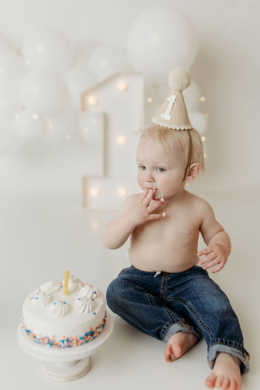 Baby in birthday hat sits beside white cake with candle, tasting frosting at first birthday cake smash photo session.