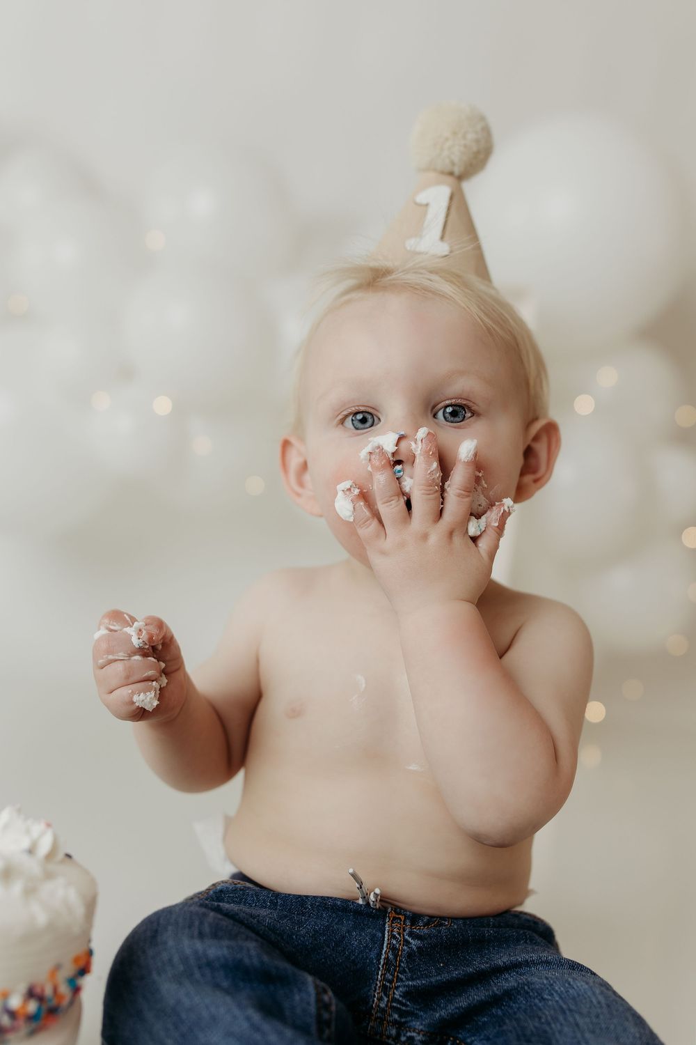 Adorable baby in party hat smashes first birthday cake, hands covered in white frosting during cake smash photoshoot.