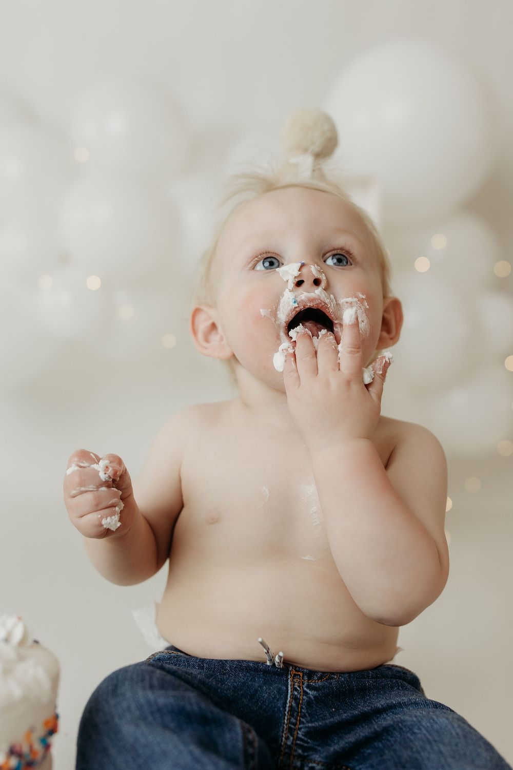 Messy baby with frosting on face enjoying first birthday cake smash in jeans with white balloons backdrop.