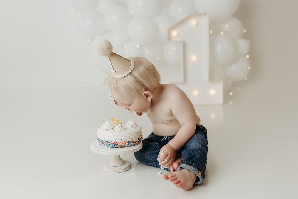 Blonde toddler in a party hat leaning over a white sprinkle cake on his first birthday.