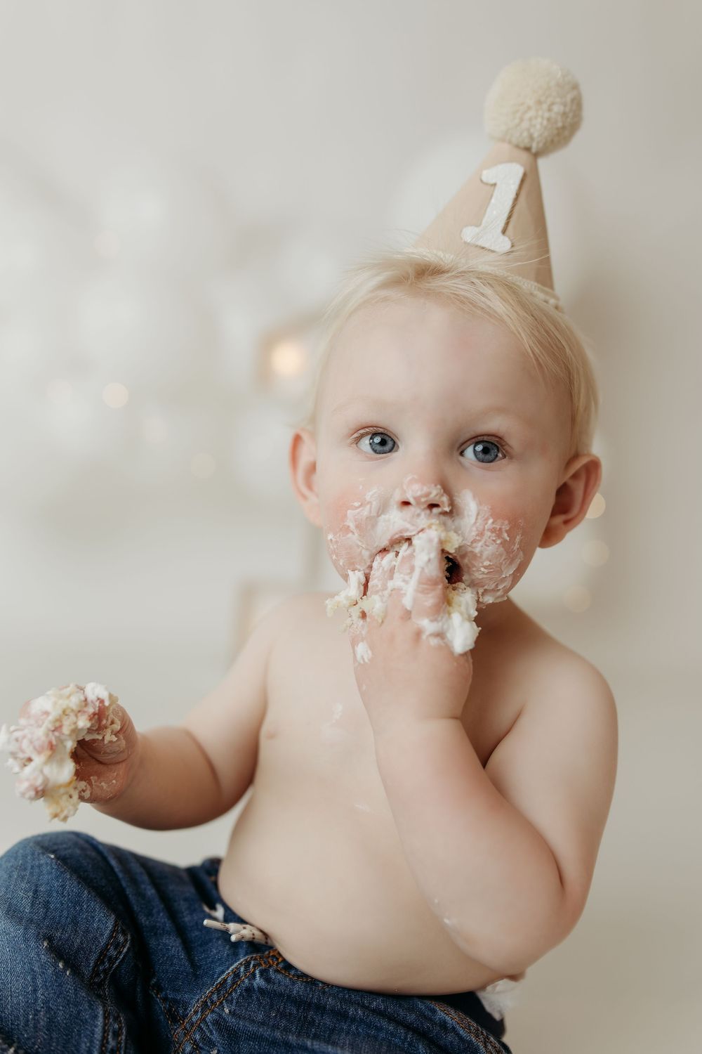 Baby's first birthday cake smash with frosting on face, wearing party hat with number 1, jeans, light background.