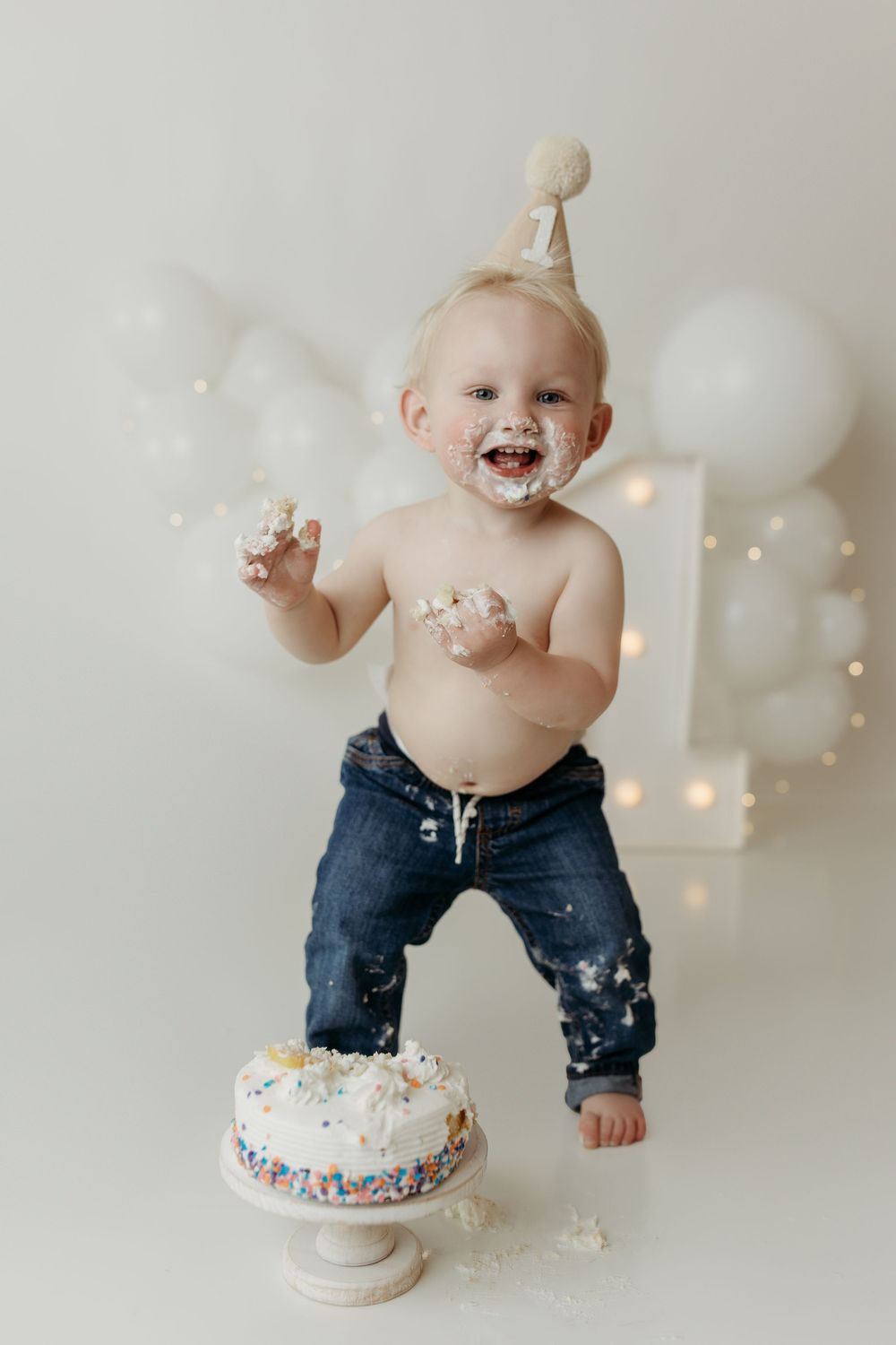 Happy toddler in party hat smashing first birthday cake with frosting covered hands and white balloon decor.