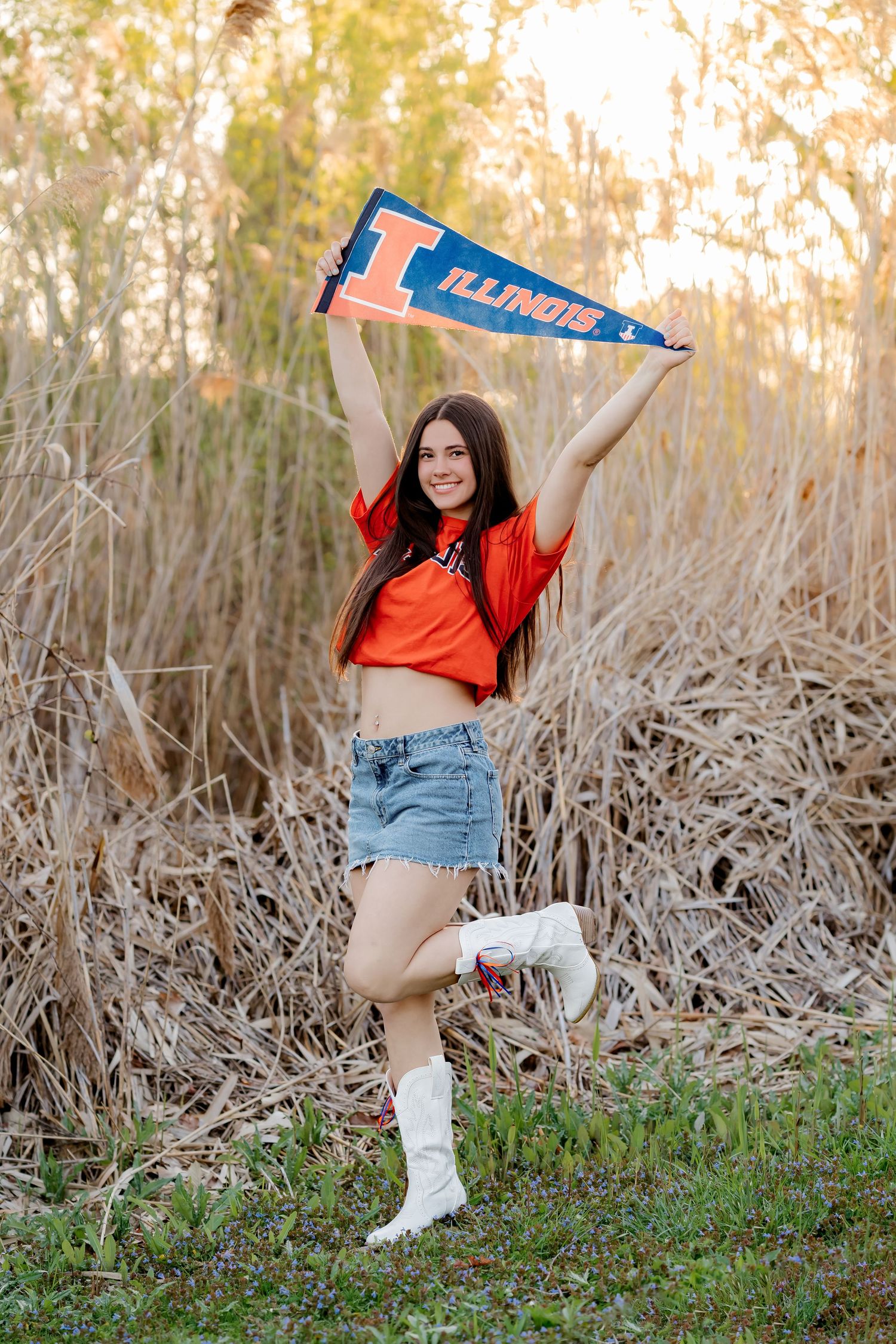Young woman in orange Illinois crop top and denim shorts holds up an Illinois pennant flag outdoors.