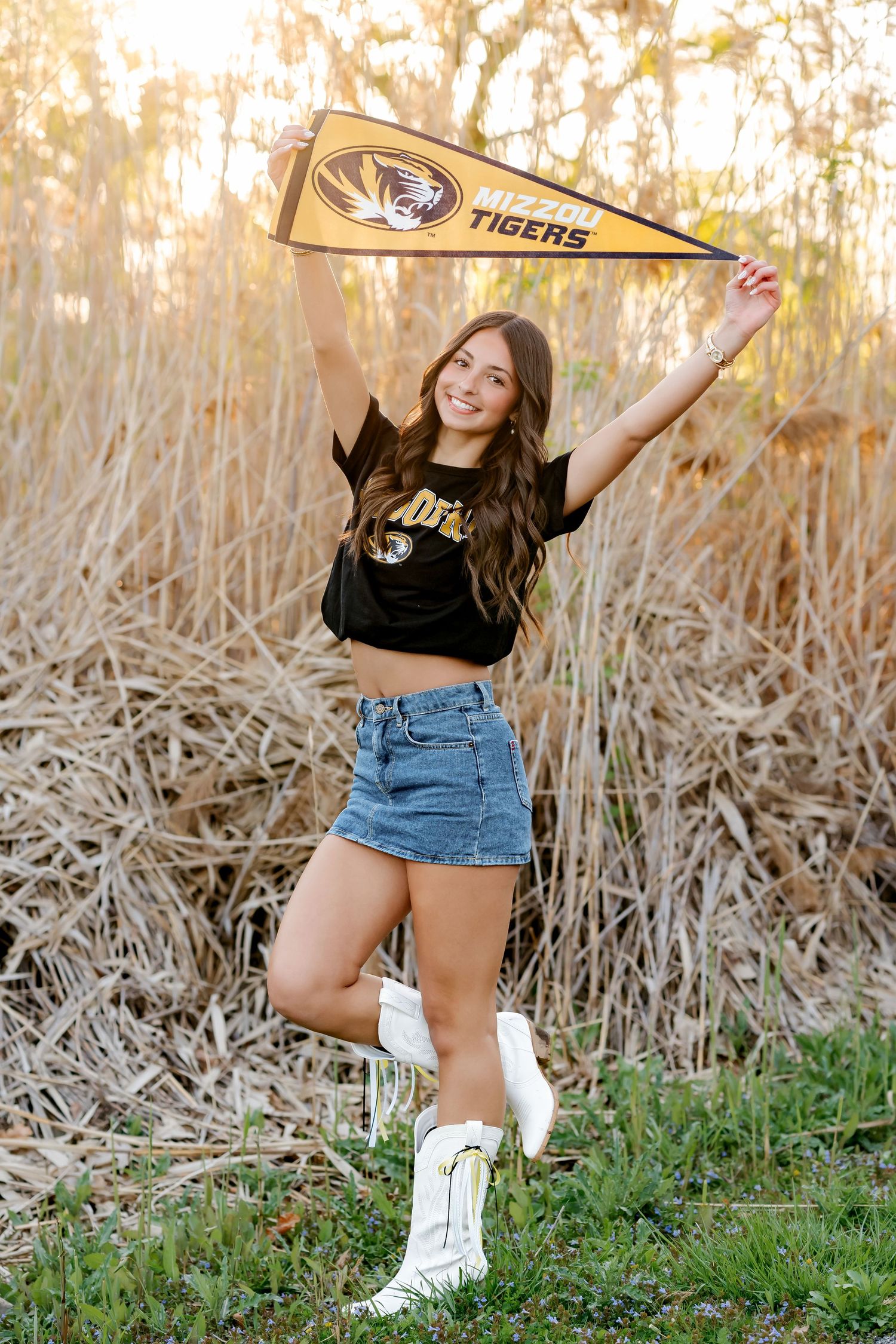 Smiling girl in Mizzou Tigers crop top and denim skirt holds pennant flag in white cowboy boots outdoors.