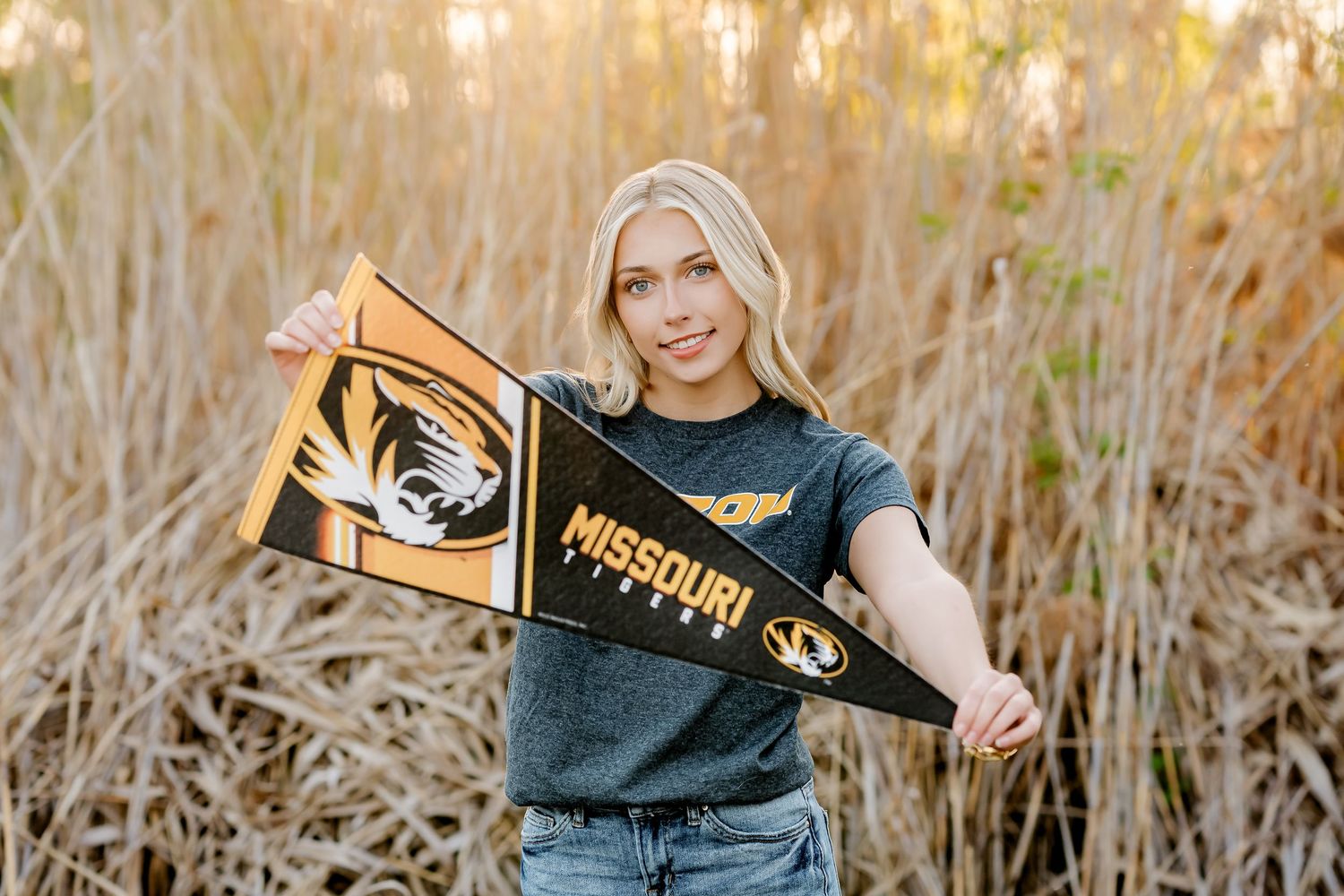 Smiling blonde student holding University of Missouri pennant flags outdoors among tall golden grasses.