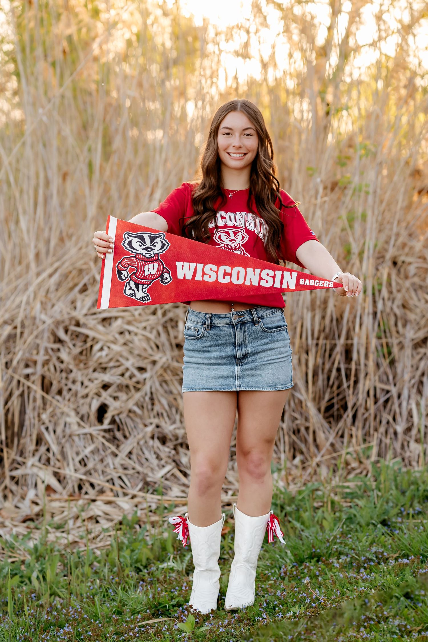Smiling girl in Wisconsin Badgers red shirt and denim skirt holds a Wisconsin pennant outdoors.