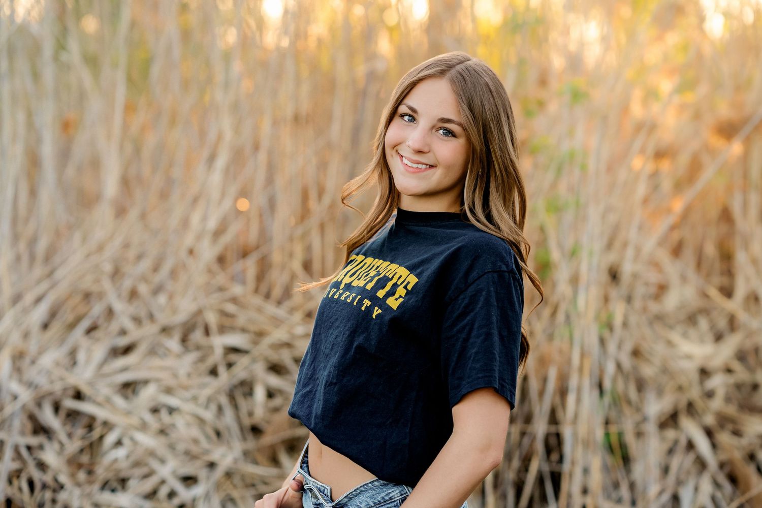 Smiling teenage girl in navy university t-shirt poses outdoors among golden dried reeds for senior portrait.
