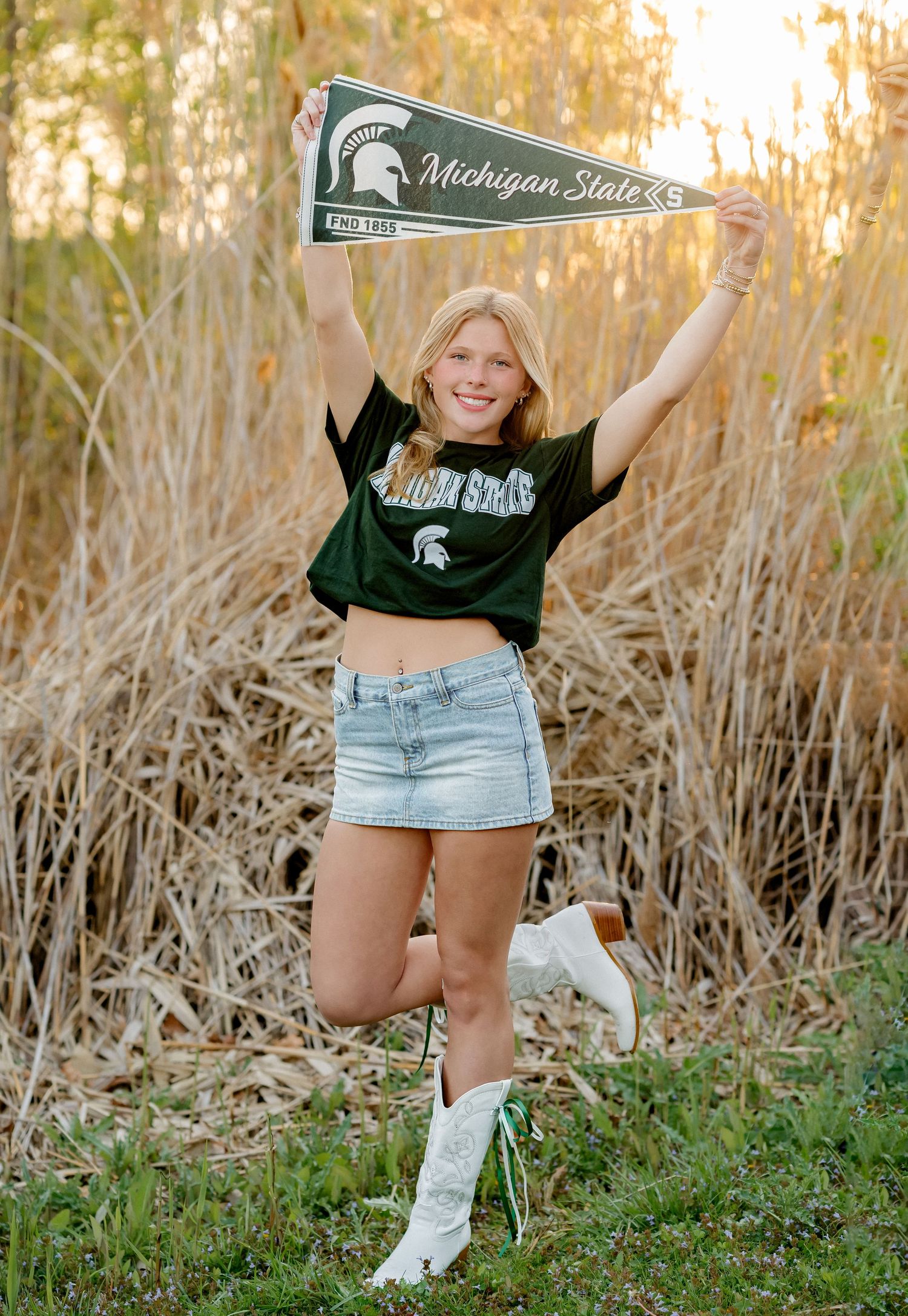 Blonde girl in Michigan State crop top and denim skirt holds pennant flag wearing white cowboy boots outside.
