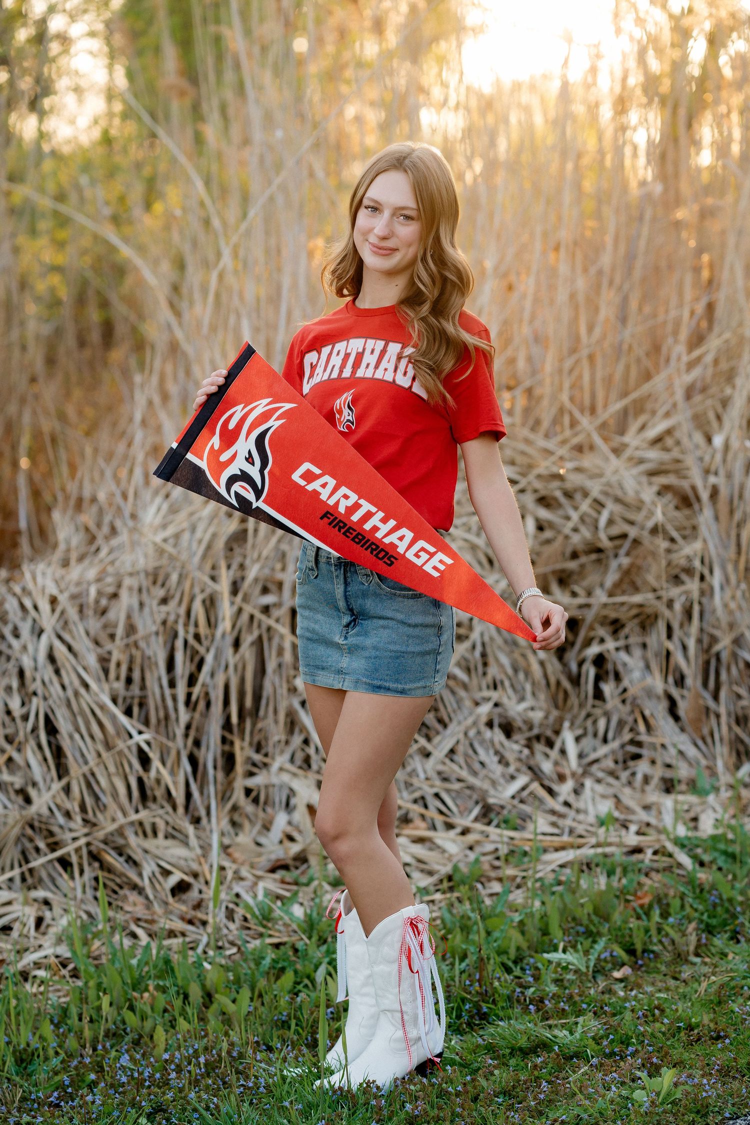 Young woman in Carthage red shirt and denim skirt holds a Carthage pennant in a natural setting.