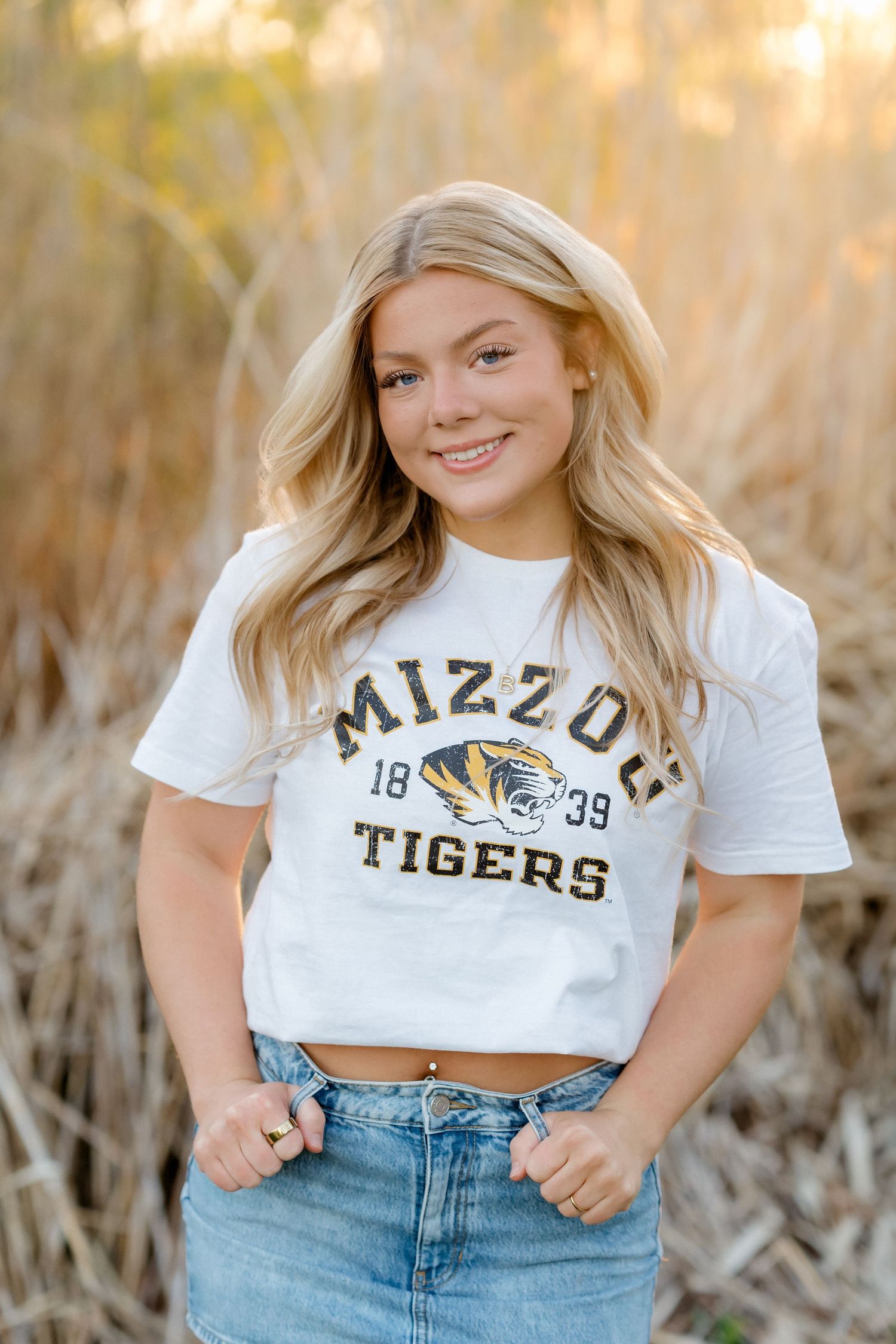 Blonde girl smiling in a white Mizzou Tigers crop tee and denim jeans outdoors.