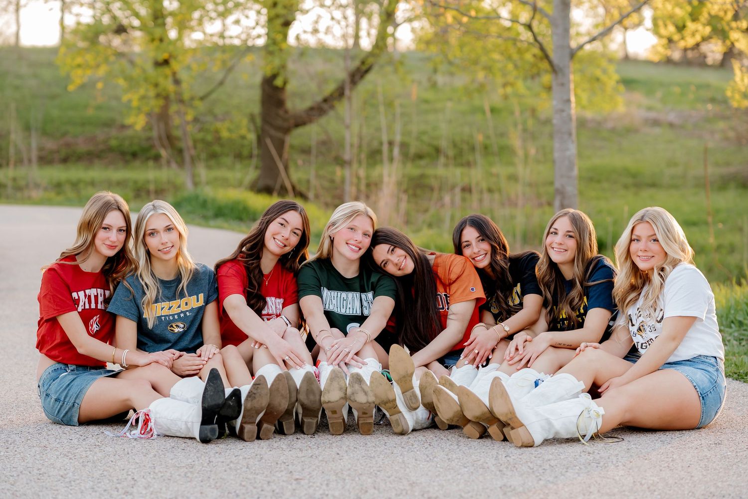 Eight teenage girls sitting together on a path outdoors, wearing college shirts and denim shorts, smiling for a group photo.