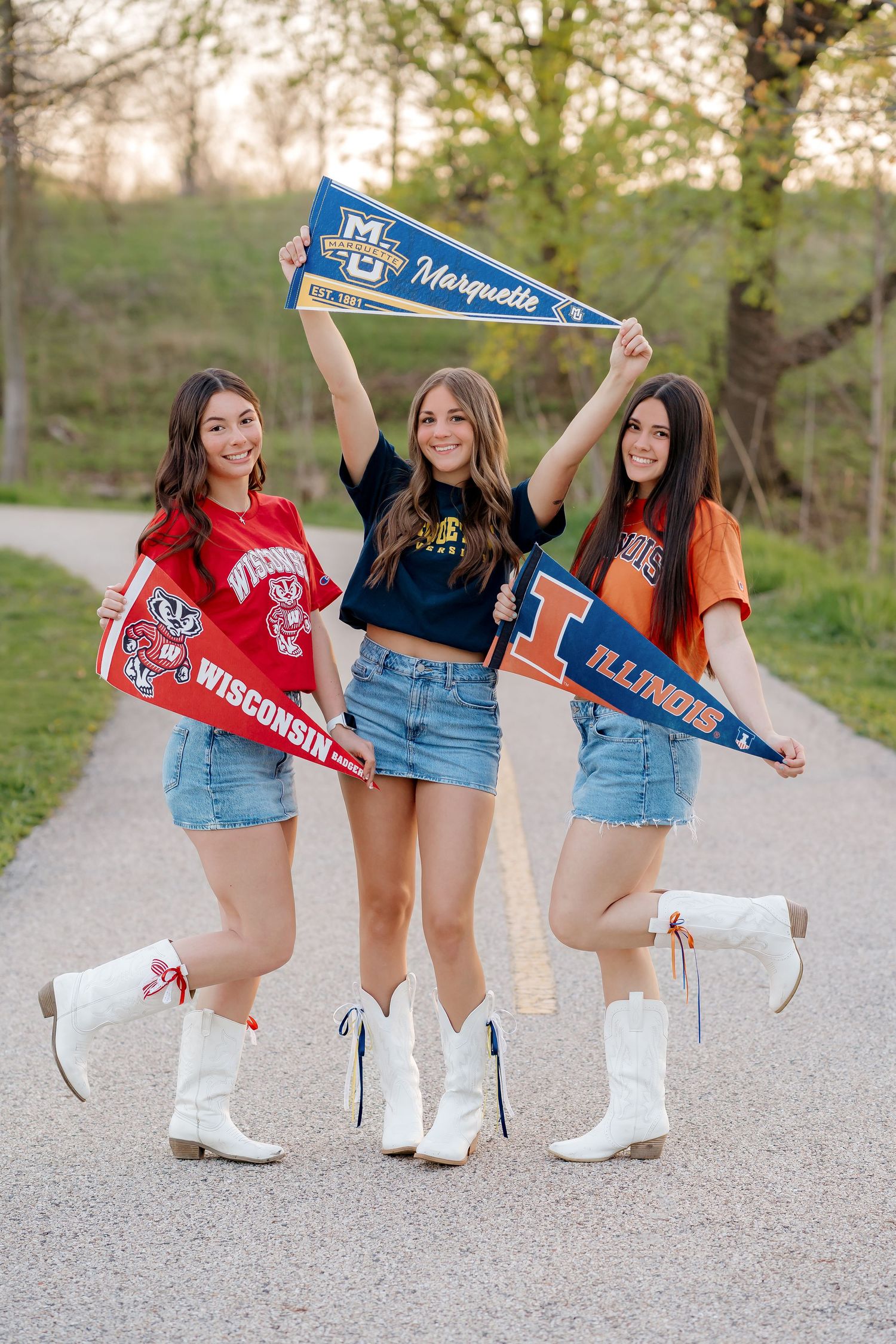 Three girls in college gear hold Wisconsin, Marquette, and Illinois pennants on a path, wearing white cowboy boots.