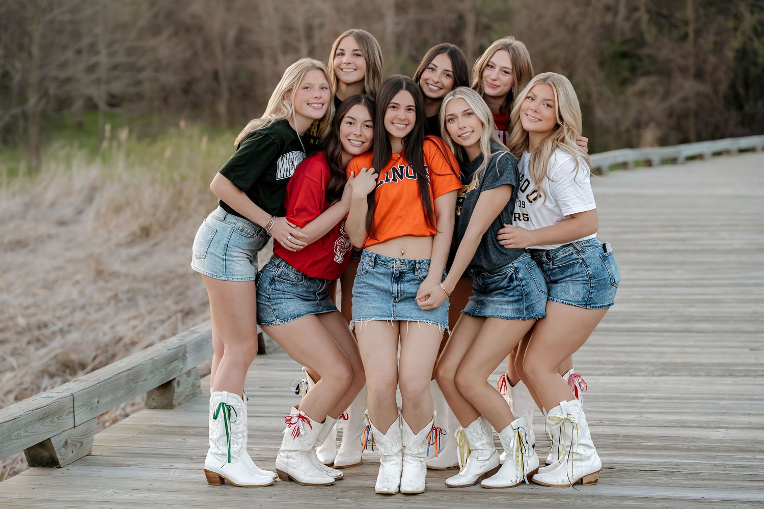 Group of nine young women posing together on a wooden boardwalk, wearing colorful tops, denim shorts, and white boots.