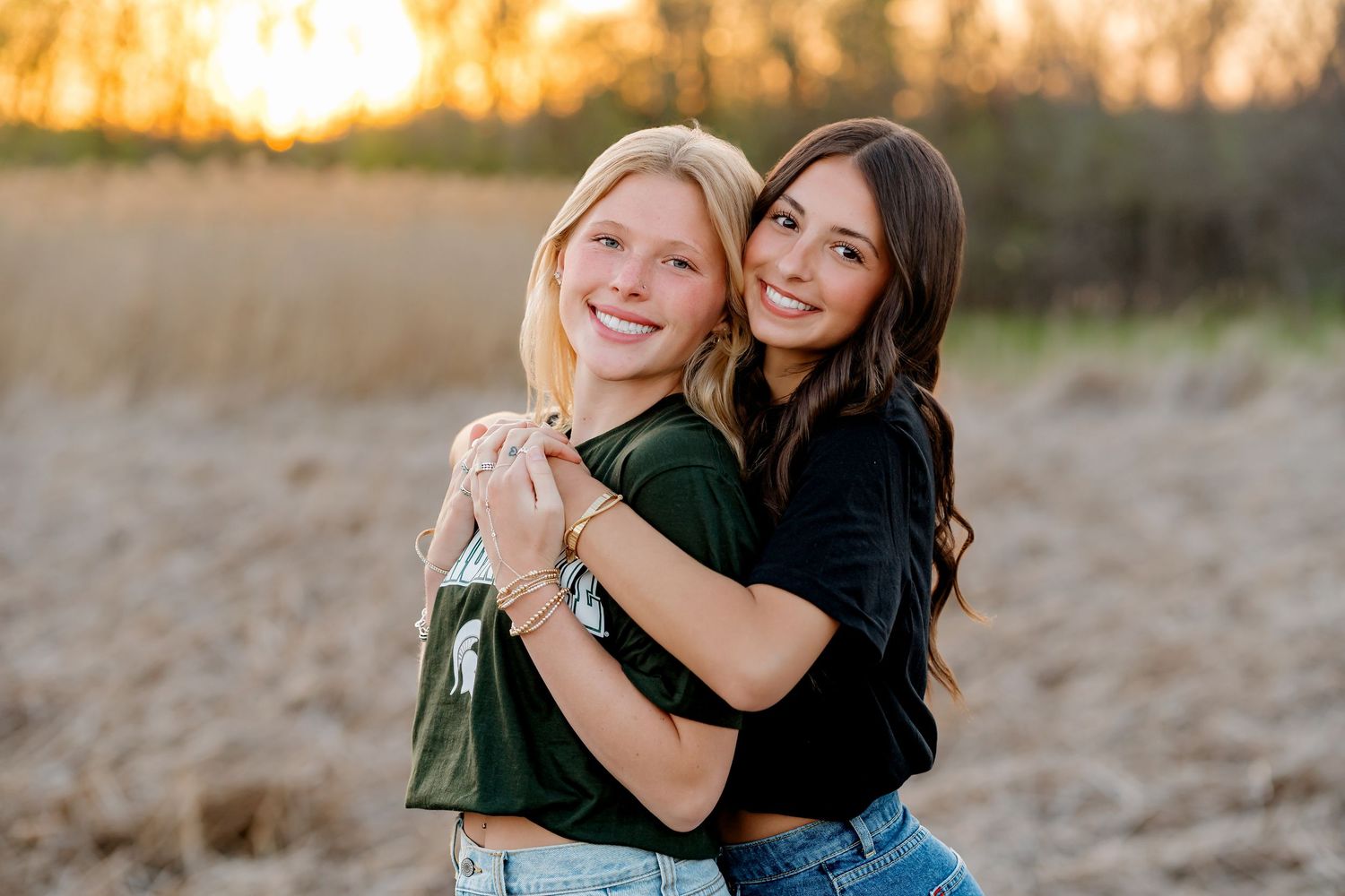 Two young women hugging and smiling outdoors at golden hour in a field, one blonde in green, one brunette in black.