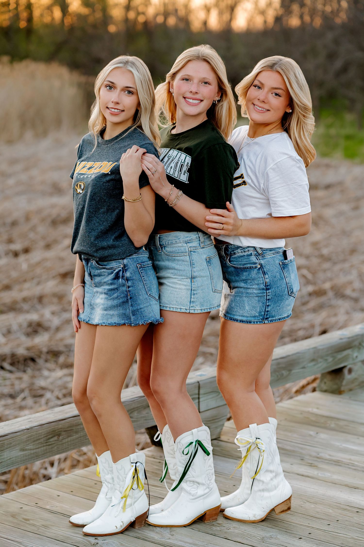 Three blonde teenage girls in denim skirts and white cowboy boots pose together on a wooden deck at sunset.