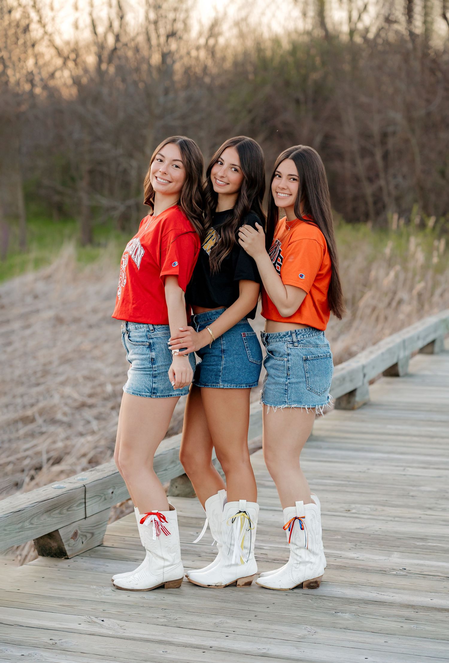 Three girls in colorful college shirts, denim skirts, and white cowboy boots pose on a wooden boardwalk.