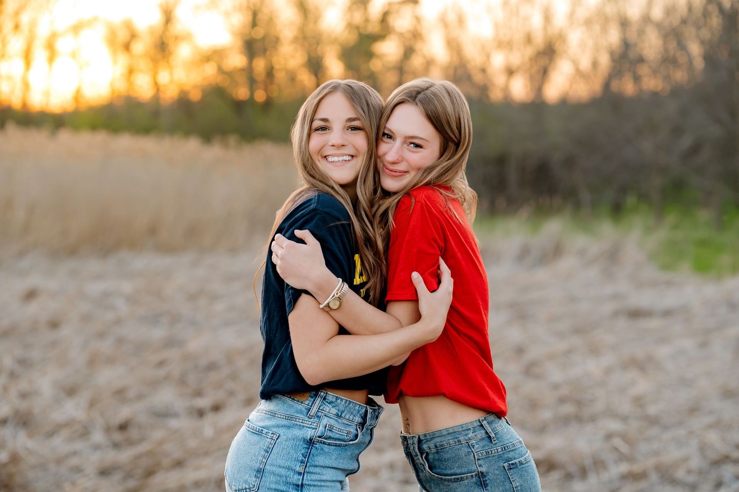 Two smiling young women hugging outdoors in a golden field at sunset, wearing casual jeans and colorful tops.