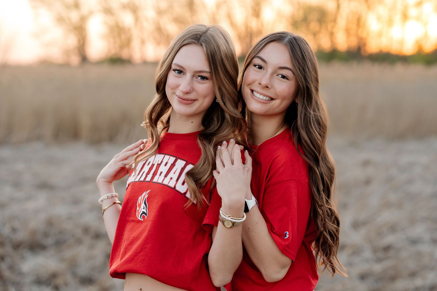 Two smiling teenage girls in matching red cardinal shirts pose together outdoors at golden hour.