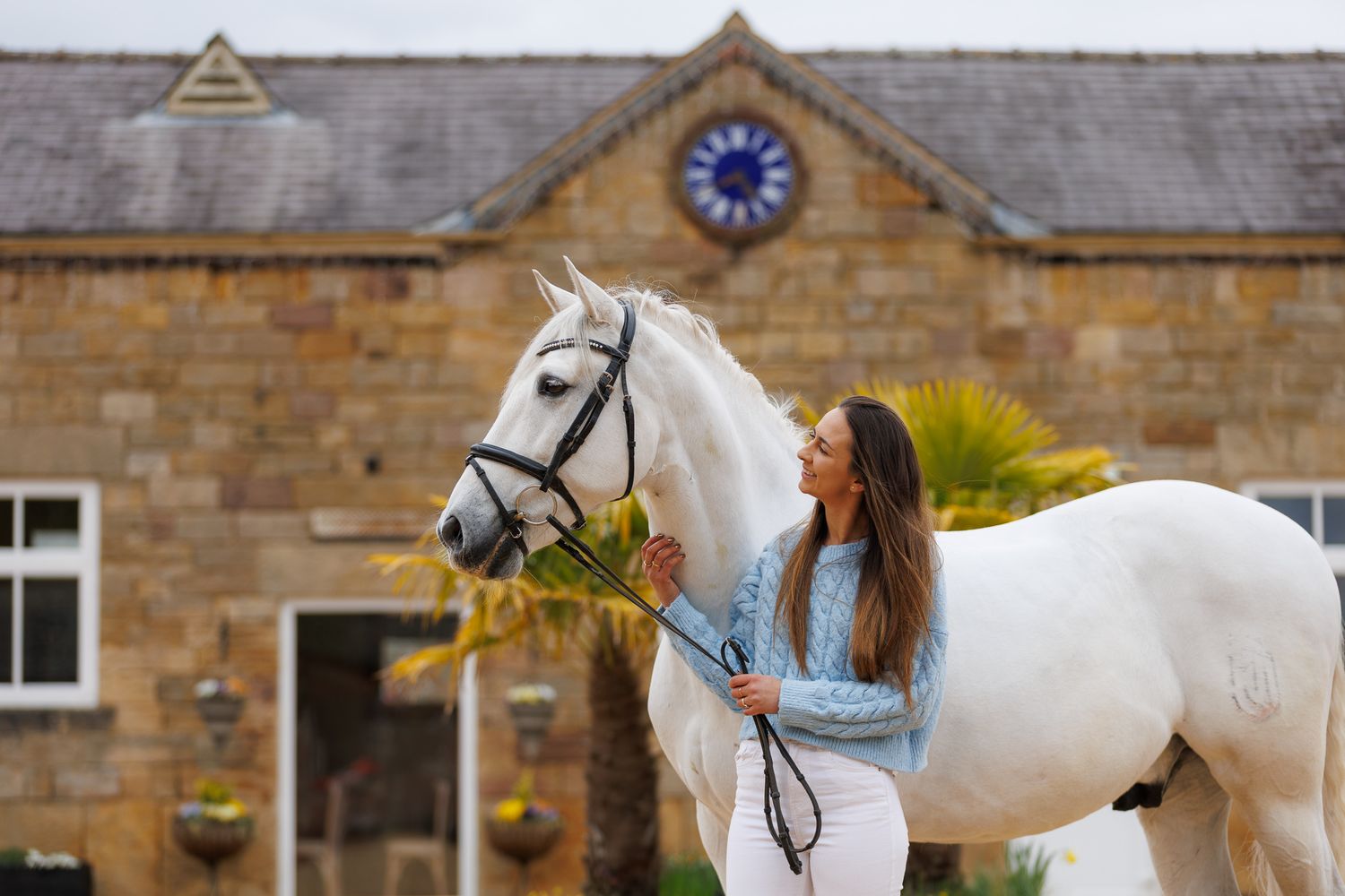 Young girl standing between a chestnut and grey horse near ornate stone gates in a lush green estate.
